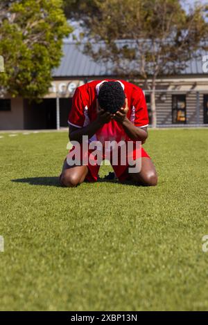 African American soccer player kneeling on grass field, appearing emotional, copy space Stock Photo