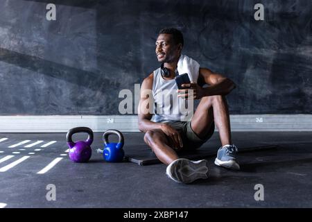 Fit African American man sitting on gym floor with smartphone after exercising, copy space Stock Photo