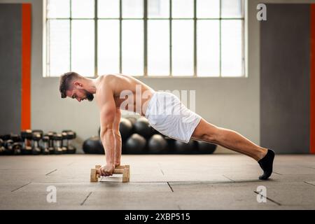 Side view of muscular male in shorts performing push up high position calisthenics exercise with handstand blocks at gym studio Stock Photo