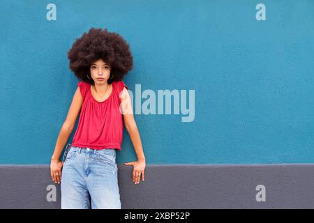Confident young Moroccan female with Afro hair looking at camera while standing and leaning with hands on wall against blue background in daylight Stock Photo