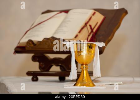 Golden chalice standing on a white altar cloth with an open bible on a wooden lectern in the background Stock Photo