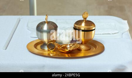 Golden tray holding religious items for a baptism ceremony, including a silver and a golden chalice, a golden shell and a white candle Stock Photo