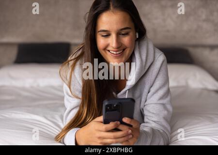 Smiling Caucasian woman in leisurewear relaxing at home, smiling while using her smartphone Stock Photo