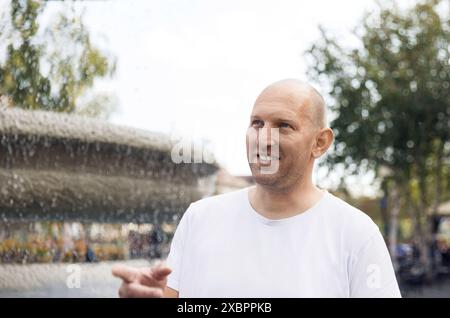 Bald man in white t-shirt smiling, pointing near fountain, outdoor park setting Stock Photo