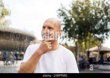 Bald man in white t-shirt touching chin, looking thoughtful, near fountain, outdoor park Stock Photo