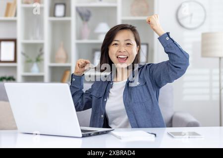 A cheerful woman expressing joy and excitement while using a laptop at home. She appears to be celebrating a successful achievement. Stock Photo