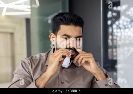 A close-up photo of a young Indian man who is being treated while in the office at the workplace, blowing his nose with medication for a sore throat. Stock Photo