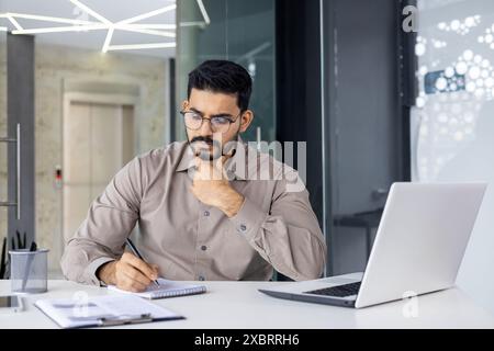 A young Indian man holds an online meeting and conference, learns remotely, sits in the office at the table, thoughtfully holds his hand by his beard, makes notes in a notebook. Stock Photo