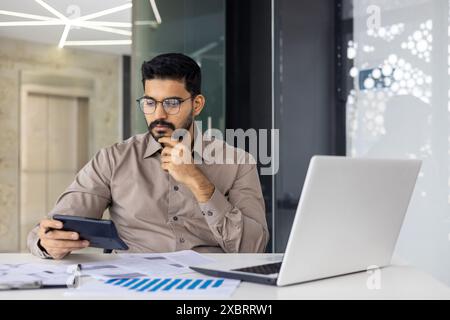 Serious and focused young Indian male accountant and expert working in office with laptop and documents, thoughtfully using calculator. Stock Photo