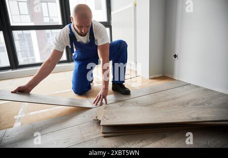 Male construction worker laying laminate wooden planks on floor underlayment. Man in work overalls installing laminate wood flooring in new flat. Home renovation concept. Stock Photo
