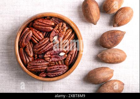 Pecan nuts in a wooden bowl on linen fabric. Dried pecan halves and whole pecans in shells, seeds and nuts of Carya illinoinensis. Stock Photo