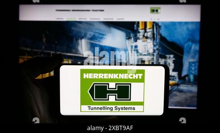 Person holding cellphone with logo of German boring machine company Herrenknecht AG in front of business webpage. Focus on phone display. Stock Photo
