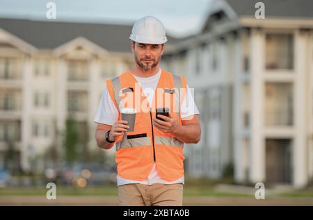 Engineer builder drinking take away coffee using phone on break. Serious engineer at new home. Construction manager in helmet. Architect at a Stock Photo