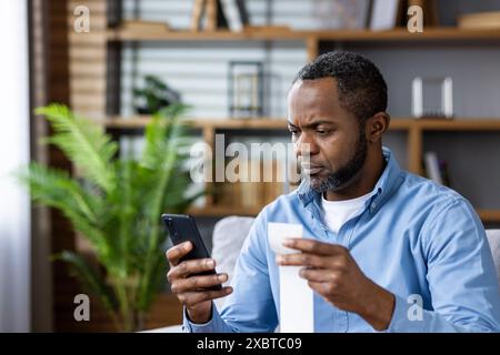 A serious man pays his utility bill for rent using an app on his phone while sitting at home. Image depicts digital payment and financial responsibility. Stock Photo