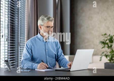 Senior man working on laptop in a modern home office. Focused and professional, he is taking notes on a notepad. Stock Photo