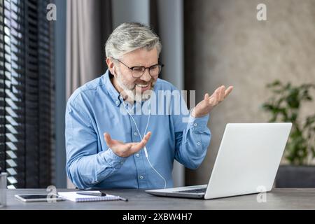 Senior man using a laptop and headset for a video call at home. He is sitting at a desk and appears to be engaged in the conversation. Stock Photo