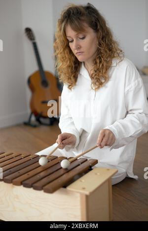 Beautiful young curly woman playing on a wooden xylophone sitting on the floor in white clothes Stock Photo