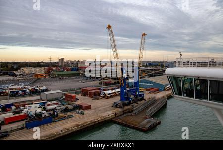 P&O cruise ship Ventura leaving Southampton showing side of the bridge with cargo containers, cranes, and trucks during dusk, with a cloudy sky Stock Photo