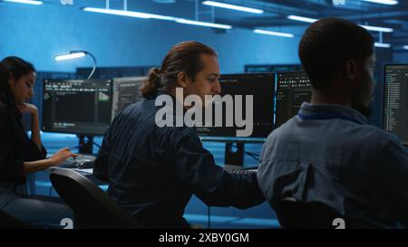 Multiracial coworkers in data center coding and checking parameters on computer. Multiethnic team working together, doing programming in high tech workspace, overseeing server clusters Stock Photo