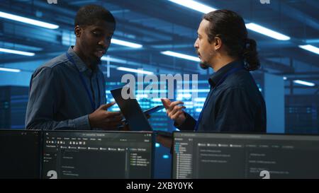 Engineers working in high tech server farm, using laptop and tablet to analyze data, ensuring seamless data flow. Men examining hardware in data center, ensuring system integrity and security Stock Photo