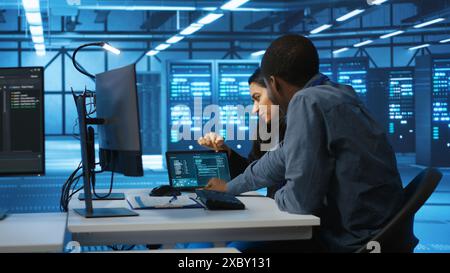 Multiethnic coworkers doing system analysis of server farm supercomputers using tablet. Diverse team of engineers looking at parameters in data center housing storage infrastructure Stock Photo