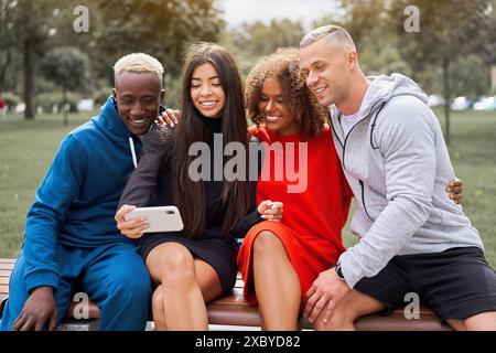 Smiling young diverse male and female friends taking selfie on smartphone. Multiethnic trendy students sitting on bench spending leisure time together in park during weekend. Stock Photo