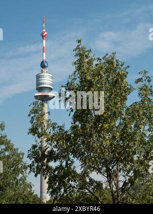 A tall television tower rises above green trees against a blue sky with some clouds, Vienna, Austria Stock Photo