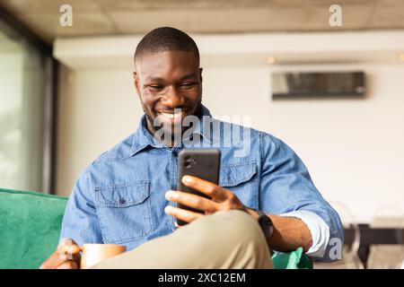 African American man smiling while looking at phone, holding cup at home Stock Photo