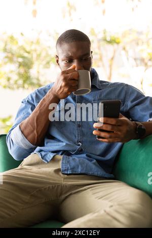 Young African American man relaxing at home, drinking coffee and using smartphone Stock Photo
