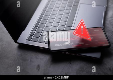 Smartphone laying on laptop with red warning notification. System warning hacked alert. Cyberattack on mobile network. Data vulnerability. Stock Photo
