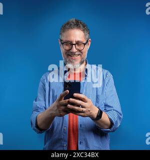 A happy senior man using a smartphone, smiling while looking at the screen on a blue background - Mature man engaging with technology - Positive and m Stock Photo