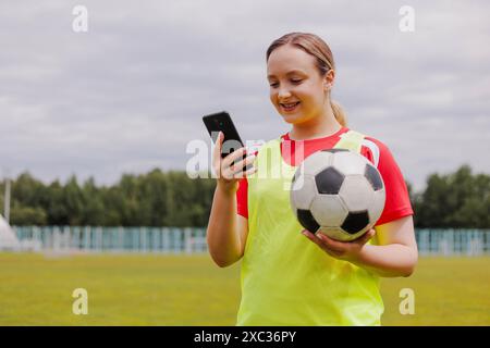 Portrait of young smiling woman football player holding soccer ball and using smartphone. Stock Photo