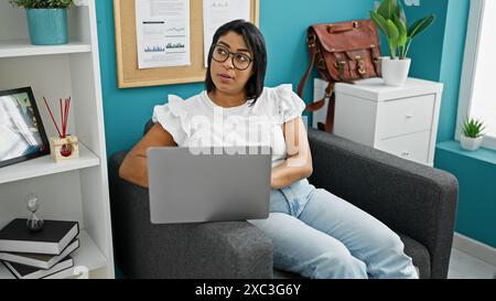 Hispanic woman working on laptop in a modern home office interior with decorative plants and shelves. Stock Photo