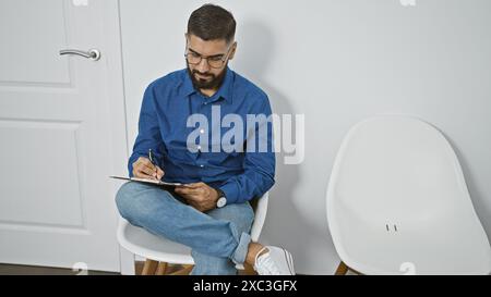 A well-groomed man with a beard sitting pensively in a white modern room, writing on a clipboard Stock Photo