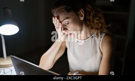 Stressed young woman experiencing a headache at night in the office, illuminated by desk lamp. Stock Photo