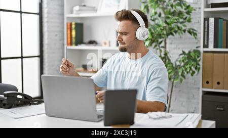 Focused hispanic man with headphones studies online in a modern home office environment. Stock Photo