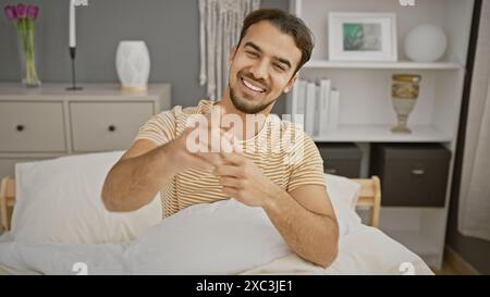 A smiling young hispanic man with a beard points at the camera in a well-lit bedroom interior, creating a relatable and inviting scene. Stock Photo