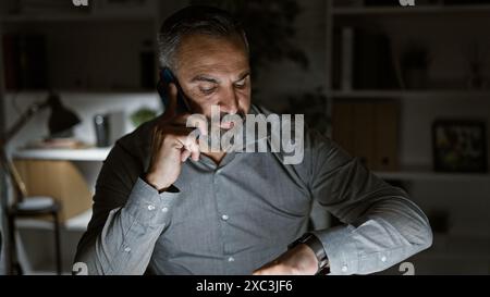 Hispanic senior man with beard in an office at night, making a call and checking the time. Stock Photo