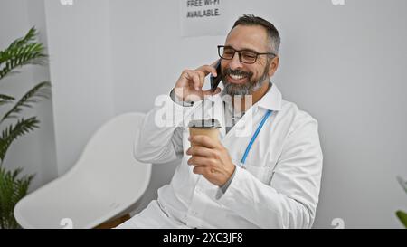 A smiling hispanic man in a white lab coat talking on a phone while holding a coffee cup in a bright clinic lobby. Stock Photo