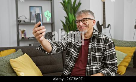 Middle-aged, grey-haired man takes selfie with phone in modern living room, exuding casual and content vibe. Stock Photo