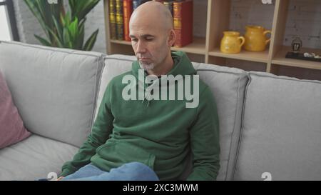 Thoughtful bald man with beard in green hoodie sitting on gray couch at home, pondering. Stock Photo
