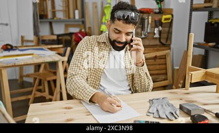 A young man talks on the phone while planning a project in his carpentry workshop. Stock Photo