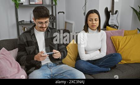 A focused man using a smartphone sits beside an annoyed woman on a couch in a cozy living room. Stock Photo