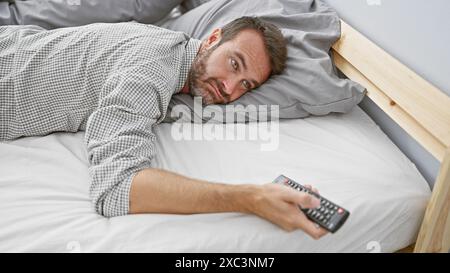 Middle-aged hispanic man with beard lying in bed holding remote control indoors. Stock Photo