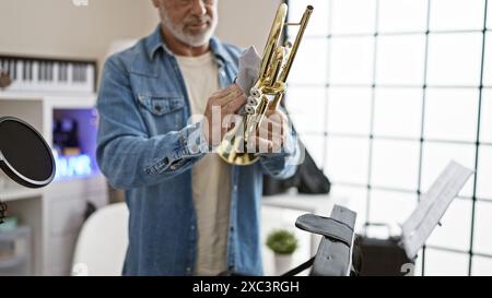 A senior man cleans a trumpet in a modern office with musical equipment. Stock Photo