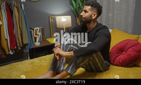 A handsome young hispanic man sits thoughtfully on a bed in a cozy bedroom, surrounded by clothes and modern decor, creating a warm and inviting home Stock Photo