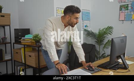 Young hispanic man working in an office environment, leaning over his desk while looking at a computer screen. Stock Photo