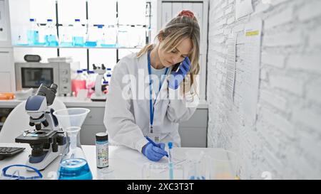 A blonde woman scientist in a lab coat analyzing a test tube while talking on the phone in a laboratory Stock Photo