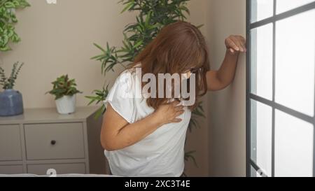 A hispanic elderly woman clutching her chest in pain stands in a cozy living room with potted plants and a window, portraying a moment of discomfort i Stock Photo