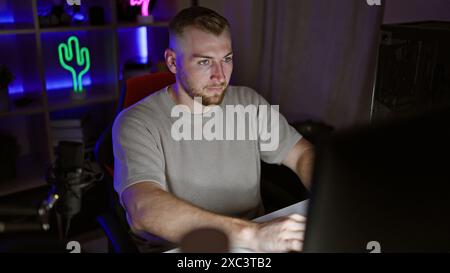 A focused young man with a beard works on a computer in a dark, neon-lit gaming room at night. Stock Photo